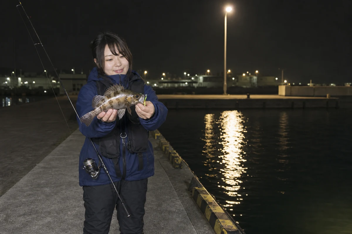 常夜灯のある夜の港で釣れたメバルを両手で見せる女性