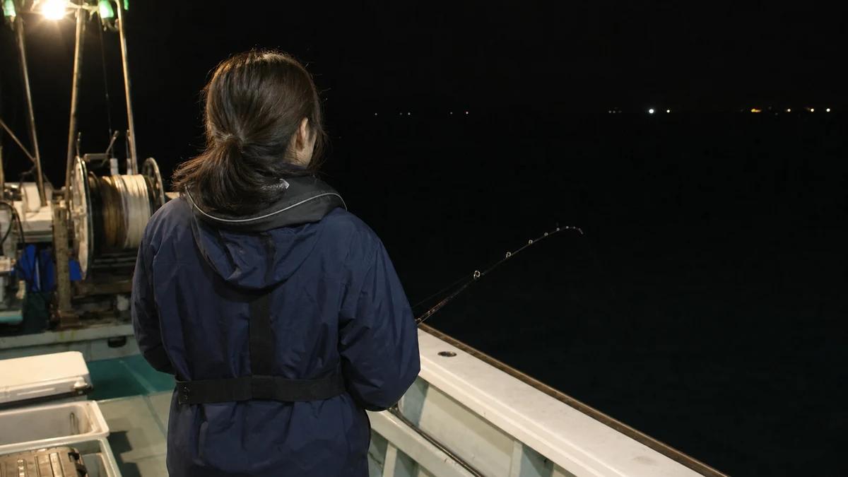 夜の船上で後ろ姿の女性がイカメタル仕掛けを指示ダナへ落としている様子